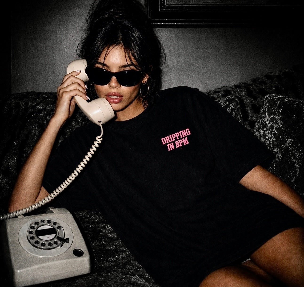 Person wearing a black t-shirt with 'DRIPPING IN 5PM' text, holding an old-fashioned phone.